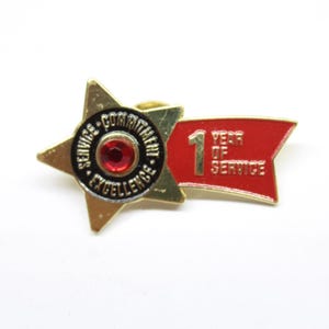 May include: Gold star pin with a red ribbon. The star has a black center with the words "Service - Commitment - Excellence" and a red jewel. The ribbon says "1 Year of Service".