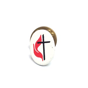 May include: An oval pin badge with a white background, displaying a black cross and a red flame emblem. The badge has a gold-coloured metal backing. The design is a symbol of the United Methodist Church.
