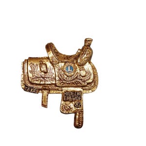 May include: Gold-colored metal saddle-shaped ornament with intricate details. The saddle has a blue and gold 'L' emblem and the text 'RIDE THE EBBTIDE 82' on the side. The ornament also has the word 'TEXAS' on the bottom.