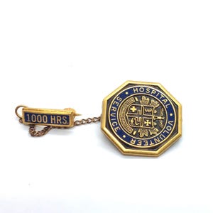 May include: Vintage gold-toned metal hospital volunteer pin with blue enamel accents. The pin features an octagonal shape with the words "HOSPITAL VOLUNTEER SERVICE" and a smaller rectangular pin that reads "1000 HRS."
