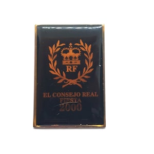 May include: Rectangular black pin with gold trim. The pin features a gold crown, laurel wreath, and the letters "RF". Below the design are the words "EL CONSEJO REAL FIESTA 2000" in gold lettering.