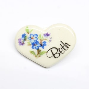 May include: White ceramic heart-shaped ornament with a blue and purple floral design and the name "Beth" written in black.