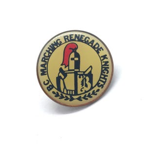 May include: A round pin with a gold border and a tan center. The pin features a knight's helmet with a red plume and the words "MARCHING RENEGADE KNIGHTS" in a circular pattern. The letters "BC" are on the left side.