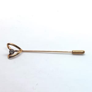 May include: A gold-colored stick pin with a heart-shaped design at the top. The heart holds a small, round, clear stone. The pin is long and slender, with a rounded end. The pin is set against a white background.