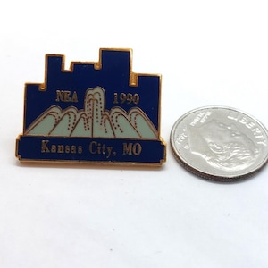 May include: A blue and gold enamel pin with the text "NEA 1990 Kansas City, MO." The pin features a cityscape design above a mountain range. A silver coin is next to the pin.