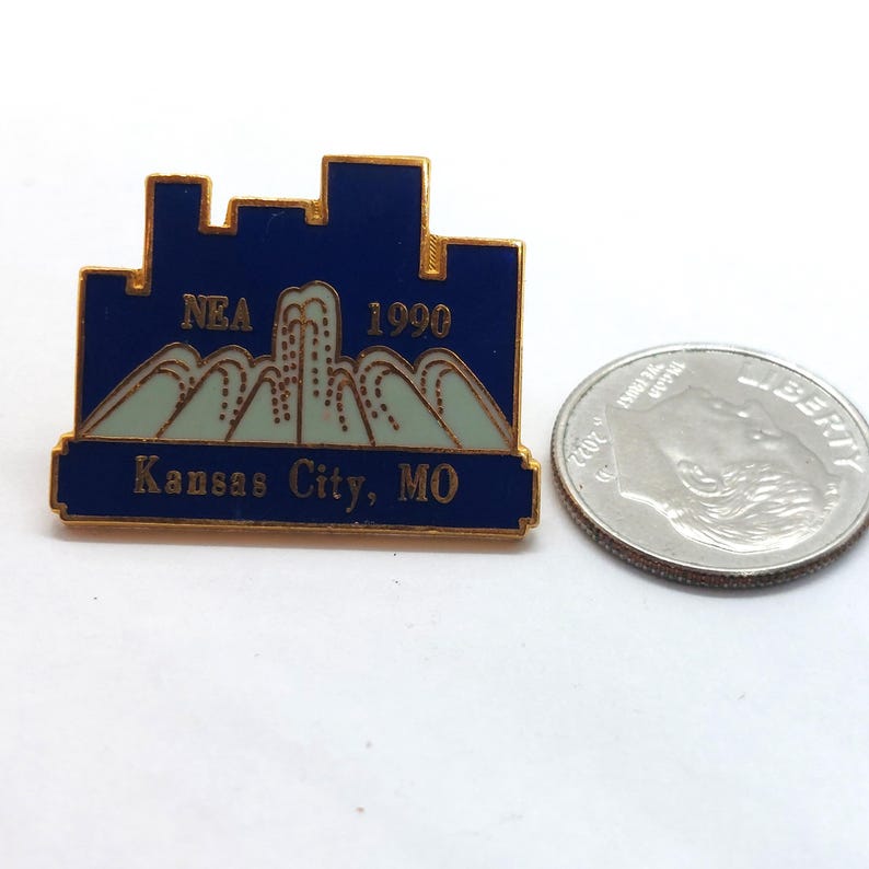 May include: A blue and gold enamel pin with the text "NEA 1990 Kansas City, MO." The pin features a cityscape design above a mountain range. A silver coin is next to the pin.