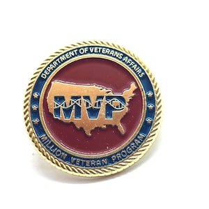 May include: A round pin with a gold border and a red center. The pin features the text "DEPARTMENT OF VETERANS AFFAIRS" and "MILLION VETERAN PROGRAM". The center has a gold outline of the United States with the letters "MVP".