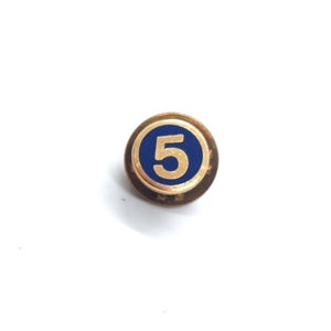 May include: A round, gold-toned pin with a blue center featuring the number 5 in gold. The pin has a simple, classic design, suitable for various uses. The pin is set against a white background.