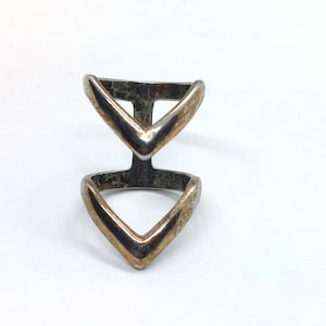 May include: A silver-toned metal ring featuring a double V-shaped design connected by a vertical bar. The ring has a polished finish with visible signs of wear and tarnishing along the edges and surfaces of the two chevron-shaped bands.