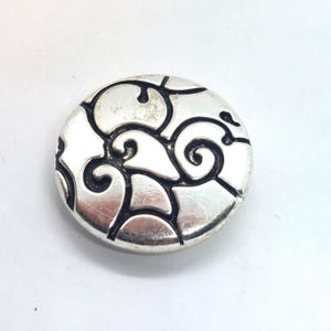 May include: A round, silver-toned button with a swirling, black, abstract design. The button has a slightly raised edge and a polished finish, creating a contrast between the silver and black elements. The design is symmetrical and detailed.