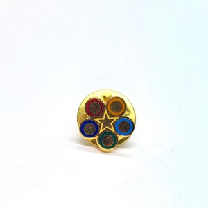 May include: A gold-colored pin featuring a star in the center, surrounded by six colorful rings. The rings are in red, orange, yellow, green, blue, and brown. The pin is circular and has a shiny finish.