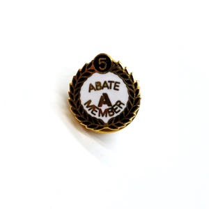 May include: A gold-colored pin with a black and white circular design. The center of the circle has the text "ABATE A MEMBER" and the number "5" in a black circle at the top.