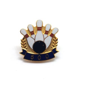May include: A gold-toned bowling pin with a black bowling ball pin. The pin features white bowling pins with red accents, a blue banner with the number "500", and a gold laurel wreath design. A sports-themed accessory.