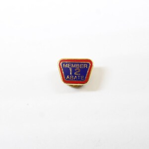 May include: A gold-toned pin with a red and blue enamel design. The pin features the text "MEMBER 12 ABATE" in gold lettering. The pin has a rectangular shape with rounded corners.