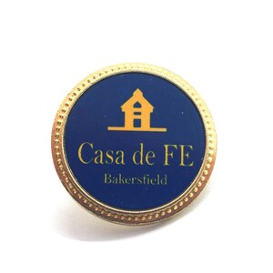 May include: A round pin with a gold-colored border and a dark blue center. The center features a gold-colored illustration of a building and the words "Casa de FE Bakersfield" in gold lettering. The pin is likely a collectible or souvenir.