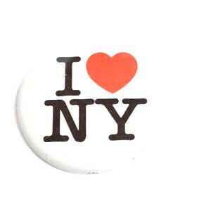 May include: White button with the text "I NY" in black letters and a red heart symbol. The button is a souvenir or promotional item, likely related to New York City.