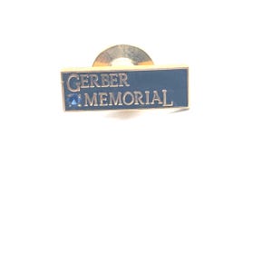 May include: A rectangular pin with the text "GERBER MEMORIAL" in gold lettering on a navy blue background. The pin has a gold-colored border and a small blue gem. The pin has a gold-colored clasp.