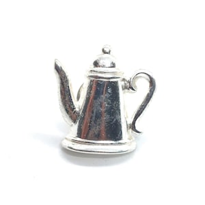 May include: A small, shiny silver-toned teapot figurine with a polished metallic finish. The object features a curved handle, a tapered spout, and a rounded lid with a small knob at the top. The surface reflects light, showing a smooth, reflective texture.