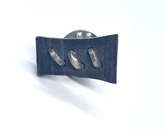 May include: A dark gray rectangular lapel pin with three silver-colored, curved accents. The pin has a silver-colored backing. The pin is set against a white background.