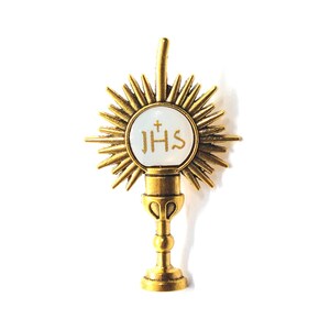 May include: A gold-colored monstrance with a sunburst design. The center features a white circle with the letters "JHS" and a cross. The monstrance has a detailed base and a vertical handle.