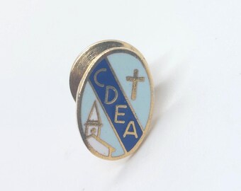May include: Gold-colored oval pin with a blue and white enamel design. The pin features a church steeple, a cross, and the letters "CDEA".