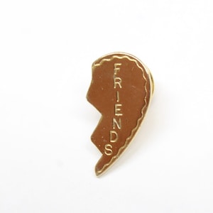 May include: A gold-colored pin shaped like half a heart. The pin has a brown center with the word "FRIENDS" vertically inscribed. The edges of the heart are scalloped, and the pin is set against a white background.