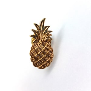 May include: A gold-toned metal lapel pin shaped like a pineapple. The pineapple features a textured, cross-hatched surface on the fruit body and detailed, pointed leaves at the top. The pin has a metallic finish with a reflective, shiny appearance.