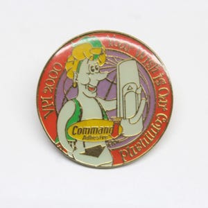 May include: A round, red, white, and purple pin with a cartoon character holding a white Command Adhesive product. The text on the pin reads "Your Wish Is Our Command" and "April 2000".