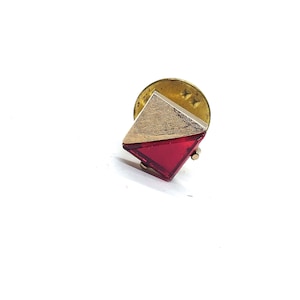 May include: A vintage lapel pin with a geometric design. The pin features a gold-toned metal square with a textured finish, and a red, diamond-shaped, transparent jewel. The pin has a round backing and a clasp.