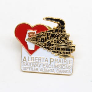 May include: A gold-coloured pin featuring a red heart and a black and gold train. The heart has a white outline of the province of Alberta. The text on the pin reads "Alberta Prairie Railway Excursions Stettler, Alberta, Canada."