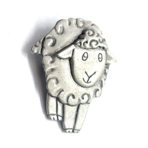 May include: A silver-colored sheep-shaped brooch. The brooch features a detailed design of a sheep with curly wool, a smiling face, and small legs. The brooch is likely made of metal and is set against a white background.