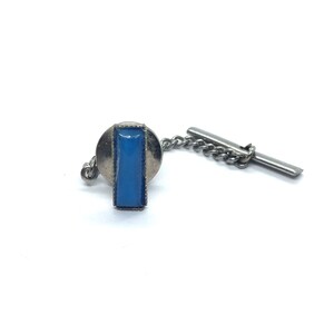May include: A silver-toned tie tack with a rectangular, light blue stone set in a silver frame. The tie tack has a chain and a bar for securing it to a garment. The background is white.