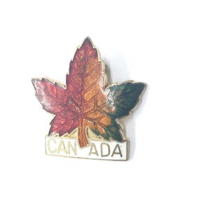 May include: A gold-colored pin with a red, orange, and green maple leaf design. The pin has the word "CANADA" written on a white banner below the leaf.
