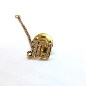 May include: A gold-colored pin with the number "10" in a textured font. The pin has a chain attached to it and a round backing. The pin is on a white background.