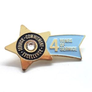 May include: A gold-colored pin with a star shape and a blue banner. The pin features the text "SERVICE COMMITMENT EXCELLENCE" around a central jewel and "4 YEARS OF SERVICE" on the banner.
