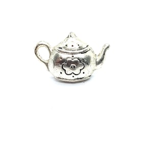 May include: A small, silver-colored teapot charm with a flower design on the front. The teapot has a handle, spout, and a lid with small holes. The charm is set against a white background.