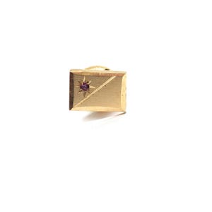 May include: A gold-colored rectangular lapel pin with a starburst design and a small, round, purple gemstone in the center. The pin has a diagonal line across the front.