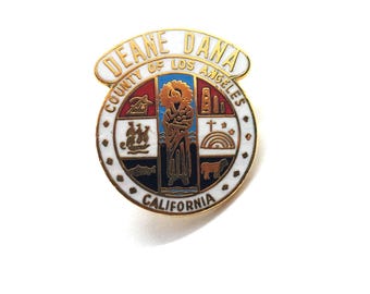 May include: A gold-toned pin featuring the seal of Deane Dana, County of Los Angeles, California. The pin has a white border with black dots and a detailed emblem with various symbols and text. The text "DEANE DANA" is at the top.