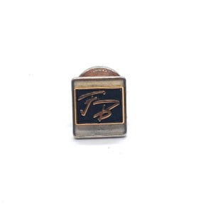 May include: A square pin with a black center and gold lettering. The pin has a silver border and a silver backing. The lettering appears to be the initials "FB" in a stylized font. The pin is on a white background.