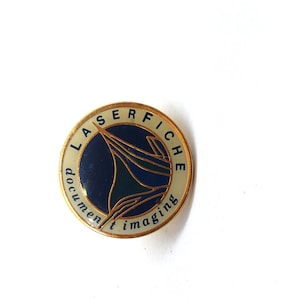 May include: A circular, gold-coloured pin featuring the text "LASERFICHE" and "document imaging". The centre has a dark blue and teal graphic. The pin is set against a white background.