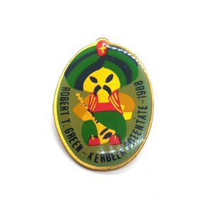 May include: Oval-shaped pin featuring a cartoon figure in a turban, holding a musical instrument. The figure is yellow with a black mustache, wearing a red vest and green pants. The pin has the text "ROBERT T. GREEN - KERBELA - POTENTATE - 1988" in green.