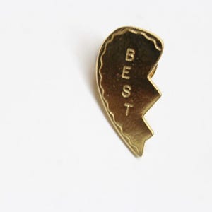 May include: Gold-colored metal pin in the shape of a broken heart. The heart has the word "BEST" inscribed on it.