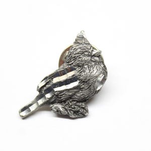 May include: A silver-toned pewter pin featuring a detailed depiction of a cardinal bird. The bird is perched with its head turned to the side, and its wings are slightly spread. The pin has a simple, classic design.