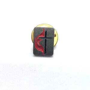 May include: A small, rectangular pin with a gold-coloured backing. The pin features a grey background with a black cross and a red flame design. The pin is a religious symbol.