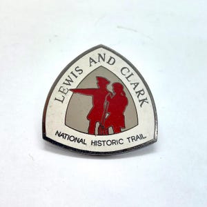 May include: A triangular metal pin featuring a white border with the black text Lewis and Clark National Historic Trail. The center displays a grey background with a red silhouette of two figures, one pointing forward and the other holding a long staff.