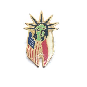 May include: A gold-tone pin featuring a depiction of the Statue of Liberty with an American flag draped behind her. The Statue of Liberty is green, black, and white. The flag is red, white, and blue.