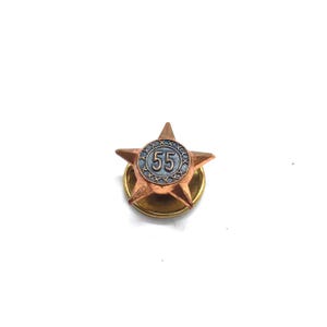 May include: A copper-toned star-shaped pin with a central circular design featuring the number 55. The pin has a gold-colored base and a classic, vintage aesthetic.