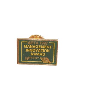 May include: Gold pin with a green rectangular background. The text on the pin reads "APTA 1997 Management Innovation Award" and "NJ TRANSIT".