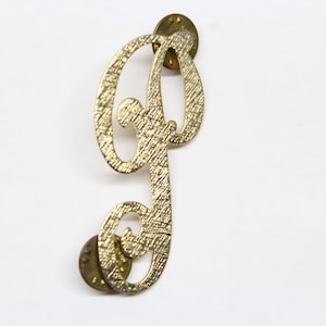 May include: Gold-toned metal letter 'P' with a textured finish. The letter is attached to a pin back.