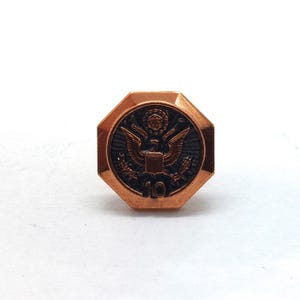 May include: An octagonal copper-toned metal pin featuring a central embossed design of an eagle with outstretched wings, a shield, and a small circular emblem above. The number 10 is clearly engraved at the bottom of the dark, recessed center.